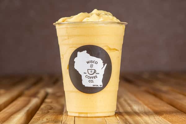 Iced mango smoothie or blended fruit frappe in a clear plastic cup with a creamy texture, topped with whipped cream, set on a rustic wooden table in a bright, modern café environment.