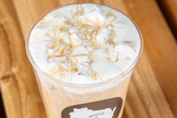 Iced white chocolate latte with espresso and milk layered over ice in a clear plastic cup, topped with light foam and a minimalist café logo, set on a rustic wooden table.