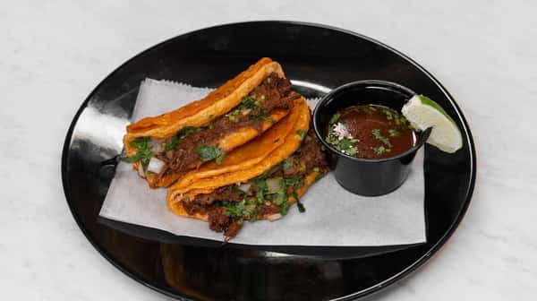 Crispy birria tacos filled with slow-cooked beef, onions, and cilantro, served with a side of savory consommé and a lime wedge on a black plate.