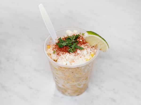 Cup of Mexican street corn topped with cotija cheese, chili powder, and chopped cilantro, garnished with a lime wedge on a marble surface.