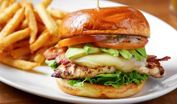 Grilled chicken sandwich stacked with melted cheese, avocado slices, crispy bacon, lettuce, and tomato on a toasted brioche bun, served with golden French fries.