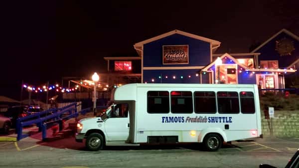 Famous Freddie's Roadhouse at night with colorful string lights glowing, and a white shuttle bus parked in front offering transportation for guests.