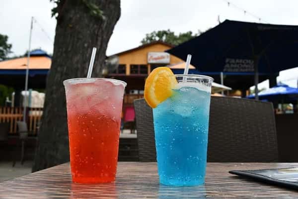Refreshing red and blue cocktails served in plastic cups with ice, one garnished with a lemon slice, sitting on a patio table at a lively outdoor bar.