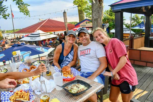 Group enjoying burgers, fries, onion rings, and drinks under patio umbrellas at a lakeside restaurant with a festive outdoor atmosphere.