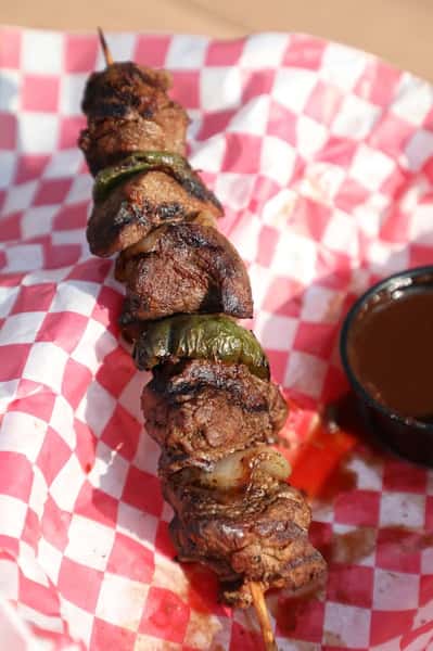 Grilled beef skewer with charred green peppers and onions, served on red-and-white checkered paper with a side of savory dipping sauce for a smoky barbecue-style dish.