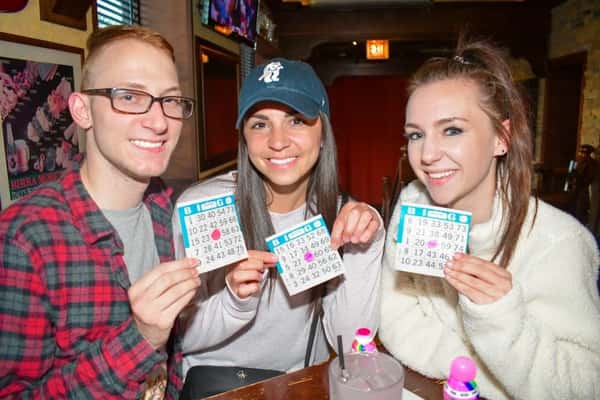 Three friends smiling and holding up bingo cards at a lively bar, with colorful bingo markers and drinks on the table, creating a fun game night atmosphere.