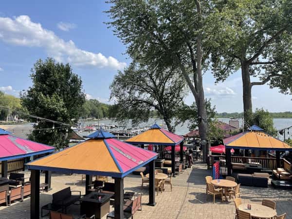 Outdoor patio seating with colorful cabana-style umbrellas, lounge furniture, and string lights, overlooking a scenic marina and lake for a vibrant waterfront dining experience.