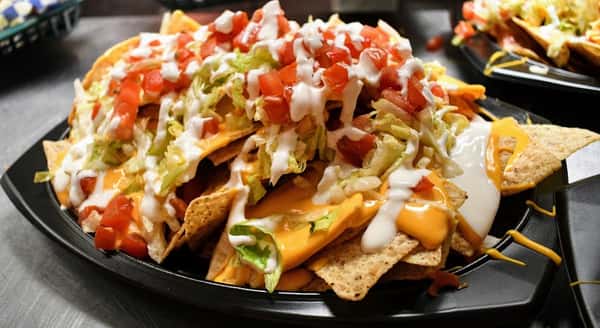 Piled-high nachos topped with melted cheddar cheese, shredded lettuce, diced tomatoes, and drizzled with sour cream, served on a black platter for a flavorful Tex-Mex appetizer.