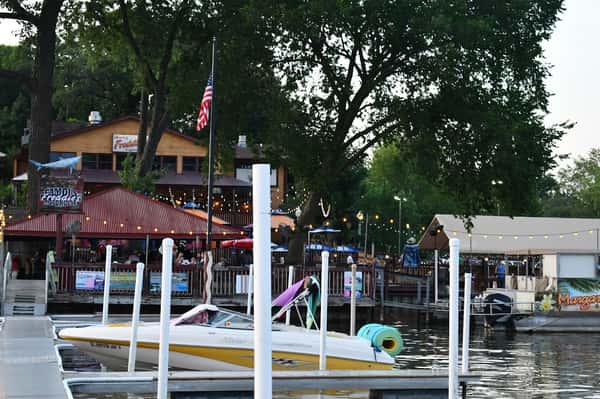 Waterfront restaurant and bar with outdoor seating, string lights, and colorful umbrellas, located along a marina with boats docked nearby, creating a lively lakeside dining atmosphere.