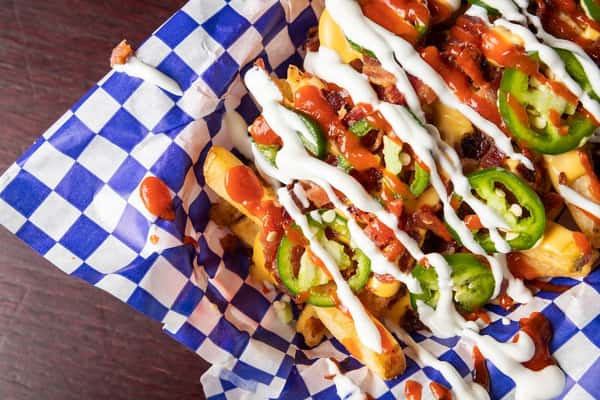 Loaded fries topped with melted cheese, crispy bacon, fresh jalapeño slices, and drizzled with ranch and hot sauce, served on blue-and-white checkered paper for a spicy and savory bar-style snack.