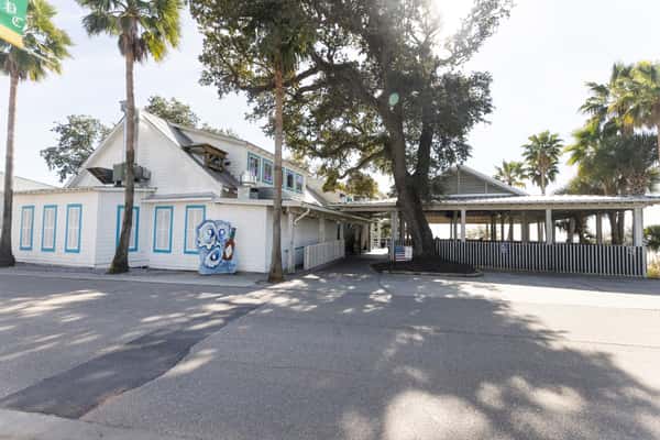 Exterior view of Bacchus restaurant with white siding, blue trim, palm trees, and covered outdoor seating in a coastal setting.
