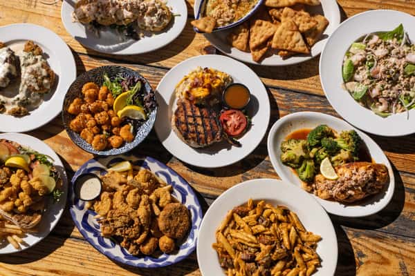 Overhead view of a rustic wooden table filled with assorted bar and grill dishes, including grilled steak with mashed potatoes, fried chicken tenders, crispy wings, loaded pasta, roasted broccoli, fresh salad, and appetizers, showcasing a hearty local American bar and grill menu.