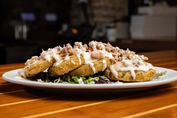 Crispy fried seafood cakes topped with lump crab meat, shrimp, and creamy sauce, served over fresh greens on a white plate, highlighting coastal bar and grill seafood appetizers.
