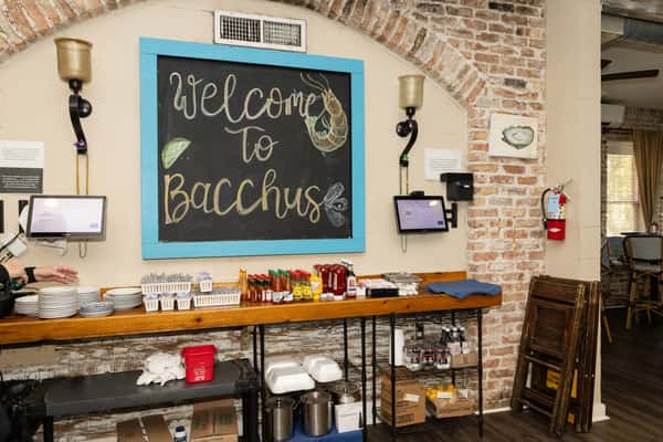 Chalkboard sign reading "Welcome to Bacchus" above a rustic host stand with hot sauces and brick walls, showcasing the warm, casual atmosphere of a coastal seafood restaurant.