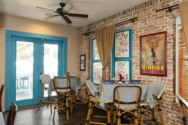 Intimate dining area with brick walls, framed artwork, patterned tablecloths, and woven chairs, emphasizing a relaxed coastal dining vibe.