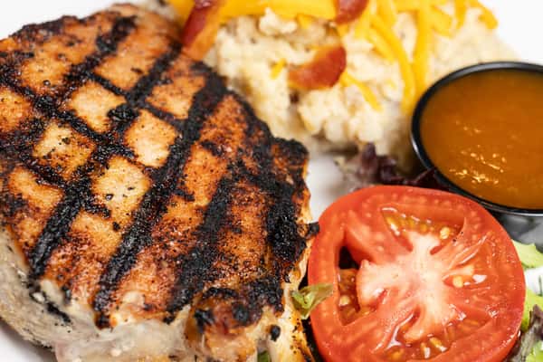 Close-up of a grilled steak with bold char marks, accompanied by mashed potatoes, brown gravy, and fresh tomato slices.