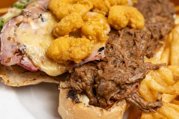 Hearty roast beef sandwich with melted cheese, bacon, crispy fried bites, and tender shredded beef on toasted bread, served with French fries, highlighting indulgent bar and grill comfort food.