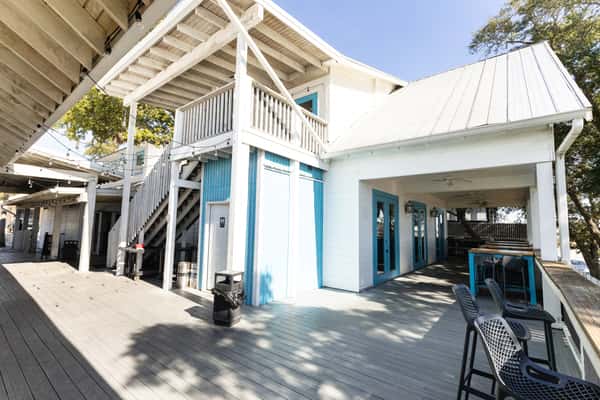Exterior view of a coastal-style bar and grill with white wood siding, blue doors, and a spacious outdoor patio deck, featuring covered seating and a relaxed, beachy atmosphere perfect for casual dining.