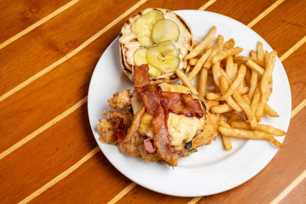 Crispy fried chicken sandwich topped with bacon and melted cheese, served with pickles and golden fries on a white plate.