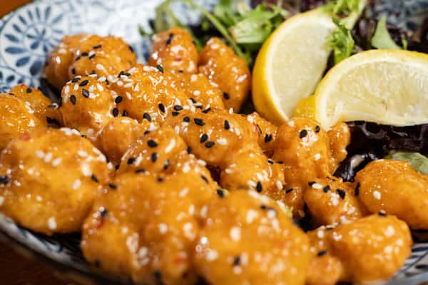 Crispy fried chicken bites tossed in a sweet and spicy sauce, topped with black and white sesame seeds, served with fresh greens and lemon wedges, highlighting Asian-inspired bar food.