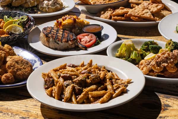 Overhead view of a rustic wooden table filled with assorted American bar and grill dishes, including grilled steak with mashed potatoes, loaded pasta, fried appetizers, roasted vegetables, and shareable plates, showcasing a hearty local bar and grill menu.