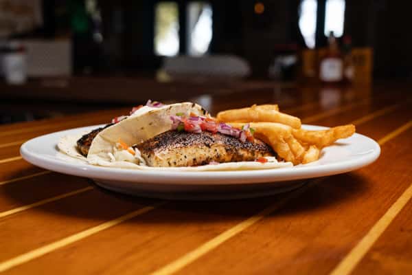 Blackened fish tacos served with fries on a white plate at a wooden bar, showcasing fresh coastal seafood cuisine.