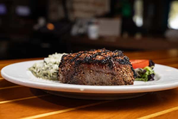 Thick-cut grilled steak seasoned with a blackened crust, served with a creamy side dish and fresh vegetables.
