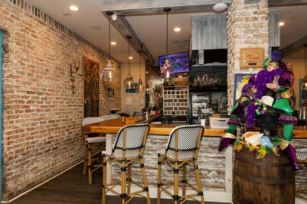 Cozy brick-walled bar interior with woven bar stools, hanging lights, and Mardi Gras décor, creating a lively coastal bar atmosphere.