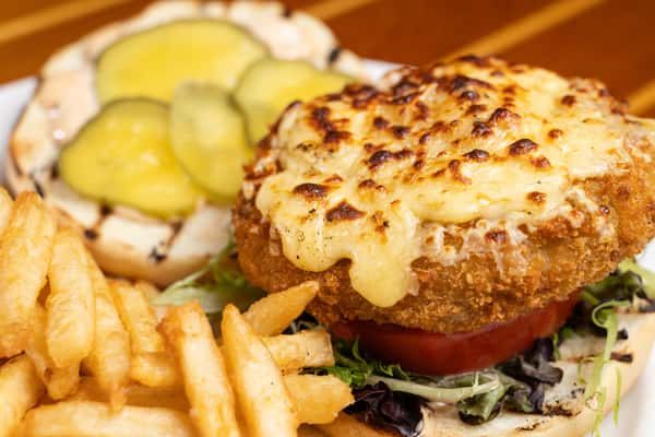 Crispy fried chicken sandwich topped with melted cheese, fresh lettuce, tomato, and pickles on a toasted bun, served with golden French fries at an American bar and grill.