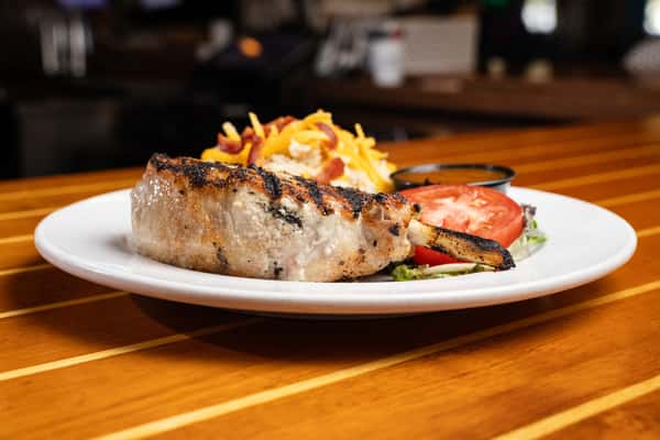 Juicy grilled pork chop with char marks, served with a loaded baked potato topped with cheddar cheese and bacon, highlighting classic American bar and grill comfort food.
