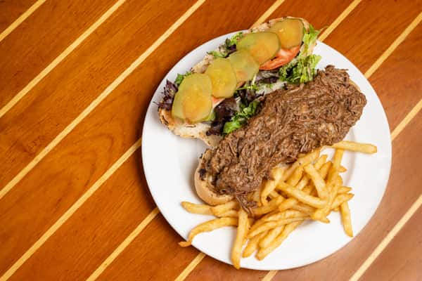 Overhead view of a roast beef sandwich with lettuce, tomato, pickles, and fries on a white plate at a casual coastal restaurant.