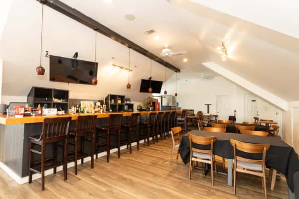 Bright bar interior with a long wooden bar, high stools, mounted TVs, and pendant lighting, creating a warm and inviting neighborhood bar and grill atmosphere.
