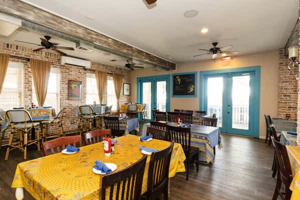 Bright dining room with brick walls, turquoise doors, ceiling fans, and patterned tablecloths, reflecting a laid-back coastal seafood restaurant ambiance.
