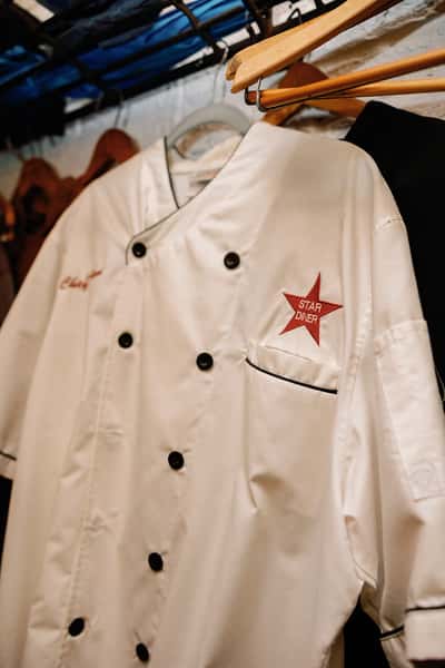 Hanging in a closet, a white chef coat belonging to the "Star Diner" is displayed. The coat features black buttons and piping, and the diner's logo, a red star, is stitched on the breast.