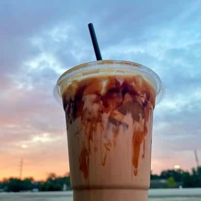 An iced drink with a sunset-colored sky in the background. The drink is drizzled with chocolate syrup inside the cup.