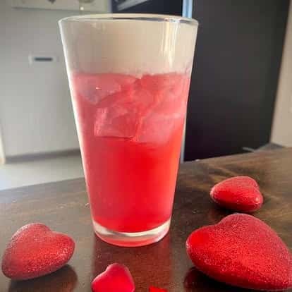 A vibrant red and clear layered drink in a tall glass, surrounded by red candy hearts.
