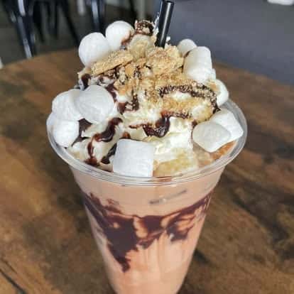 A decadent chocolate dessert drink in a clear cup, topped with whipped cream, marshmallows, chocolate drizzle, and cookie crumbs.