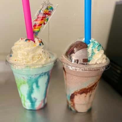 Two colorful, swirled milkshakes topped with whipped cream and sprinkles—one blue and green, the other pink and brown—served in plastic cups with thick straws.