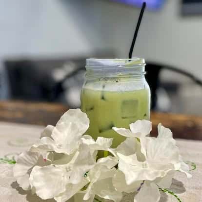 A matcha latte in a small mason jar, surrounded by delicate white flower petals on a rustic wooden table.