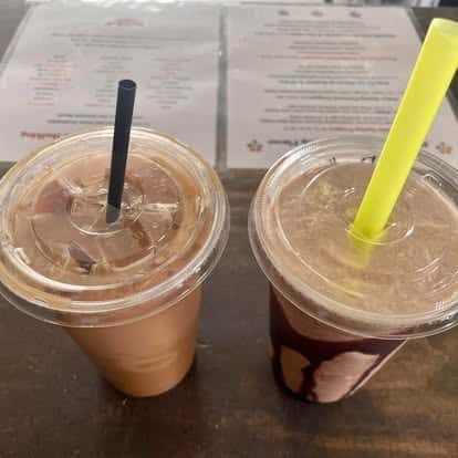 Two iced coffee drinks in plastic cups with black and yellow straws, placed on a table with a menu in the background.