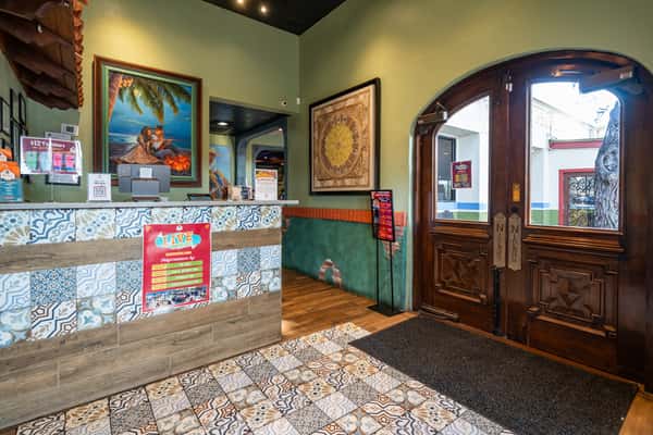 Warm and inviting restaurant foyer featuring tile-patterned hostess stand, framed artwork, and classic wooden double doors with window panes—sets the tone for an elegant Tex-Mex ambiance.