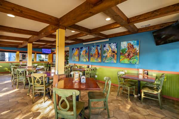 Colorful and inviting Mexican restaurant interior featuring wooden ceiling beams, green chairs, and vibrant wall art, creating a warm and festive dining atmosphere with natural sunlight and cozy seating.