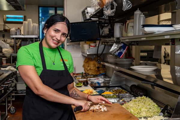 Smiling kitchen staff member preparing fresh chopped chicken at a busy restaurant prep station, surrounded by ingredients like shredded lettuce, jalapeños, and cheese.