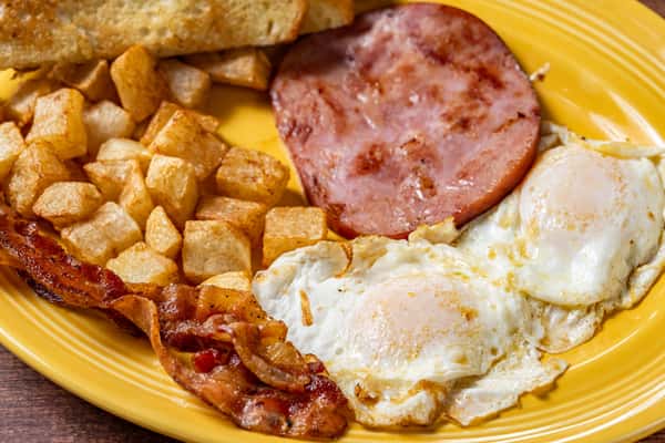 Traditional breakfast featuring bacon, hollandaise potatoes, sunny‑side‑up eggs, and a slice of grilled ham on a bright yellow plate.