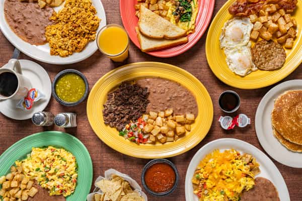 Colorful overhead spread of Mexican and American breakfast plates featuring scrambled eggs, refried beans, crispy potatoes, pancakes, chilaquiles, fresh fruit juice, salsa, and coffee, all served with toasted bread and tortillas.