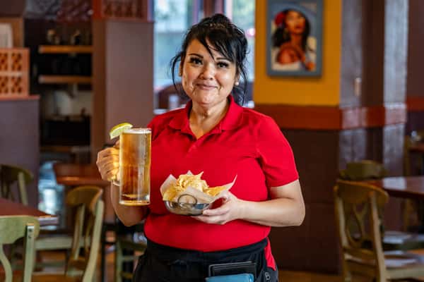 Smiling waitress in a vibrant Mexican restaurant holding a frosty mug of beer garnished with lime and a basket of tortilla chips with salsa.