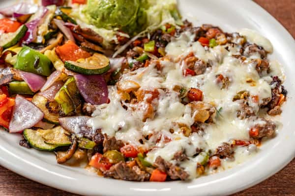 Sizzling beef fajita skillet served with grilled onions, bell peppers, zucchini, and melted cheese, topped with fresh guacamole and shredded lettuce.