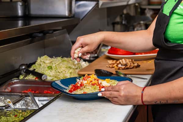 Behind-the-scenes kitchen view of a chef garnishing a fresh Mexican salad bowl with shredded cheese and diced tomatoes, surrounded by lettuce, guacamole, and grilled chicken.