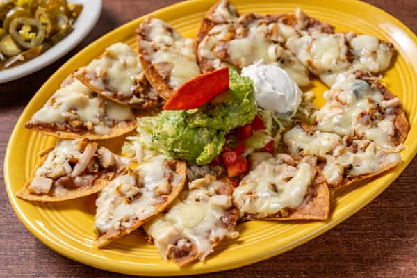Generous plate of cheesy nachos topped with shredded chicken, guacamole, sour cream, and pico de gallo, with pickled jalapeños on the side—great for sharing or appetizer feature.