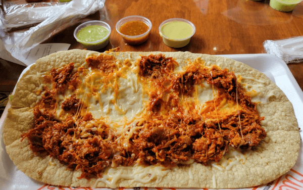 Tlayuda topped with melted cheese and seasoned shredded meat, served with three small cups of green, red, and yellow sauces.